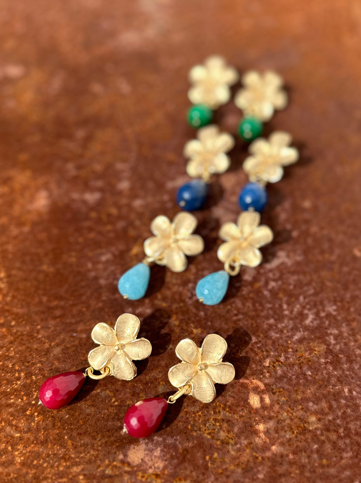 Brincos Flower dourados com pedra semipreciosa, várias cores sobre fundo terracota