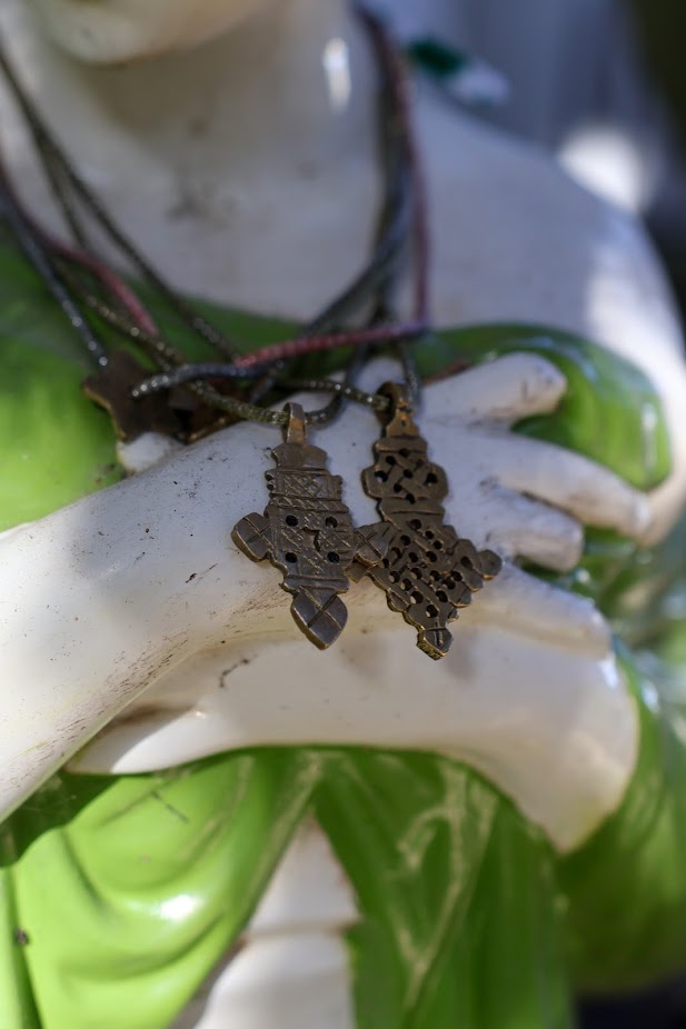 Colar Cruz Etiópia de malha metálica, dois colares na mão, sobre fundo verde